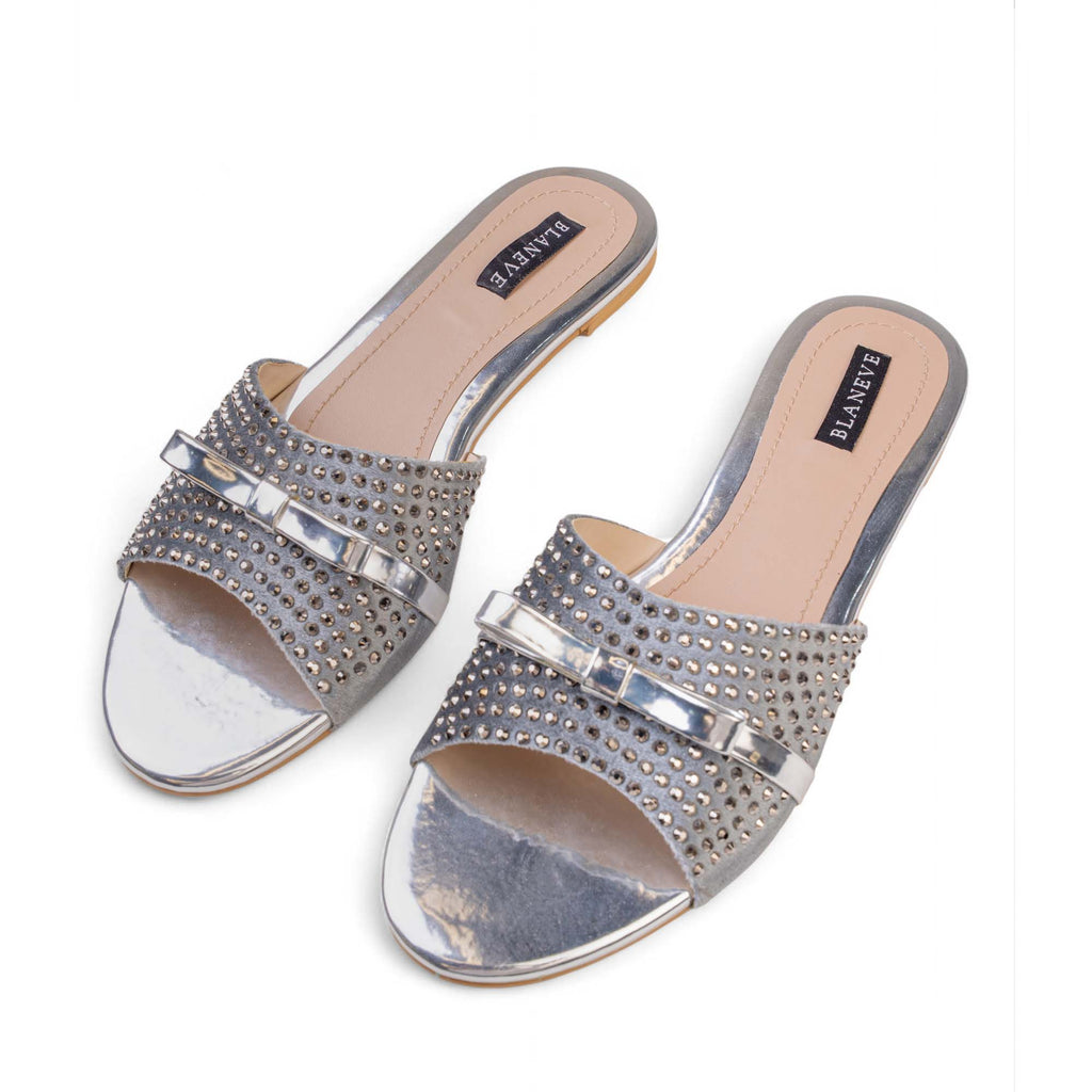 Angled view of BLANEVE Starling silver-toned slides showing crystal-covered straps and polished metallic bows.